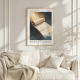 a framed poster of a roll of parchment paper hanging on a white wall above a white couch with a blanket draped over it.