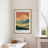 a framed poster of a mountain landscape with a person walking on a trail, hanging on a wall in a room with a white table and a window.