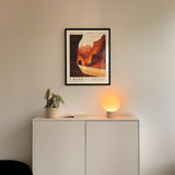 a minimalist interior design with a framed poster of a desert landscape hanging on a white wall above a white cabinet. The cabinet has a plant and a lamp on it, creating a cozy and inviting atmosphere.