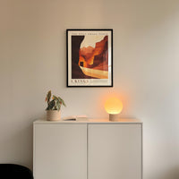 a minimalist interior design with a framed poster of a desert landscape hanging on a white wall above a white cabinet. The cabinet has a plant and a lamp on it, creating a cozy and inviting atmosphere.