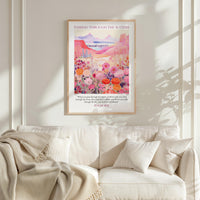 a framed poster of a vibrant, colorful landscape with flowers and mountains, hanging on a wall above a white couch with a blanket draped over it.
