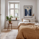 A cozy bedroom with a large bed, a nightstand with a plant, and a framed poster on the wall. The room has a warm, inviting atmosphere with a wooden floor and a window overlooking a scenic view.