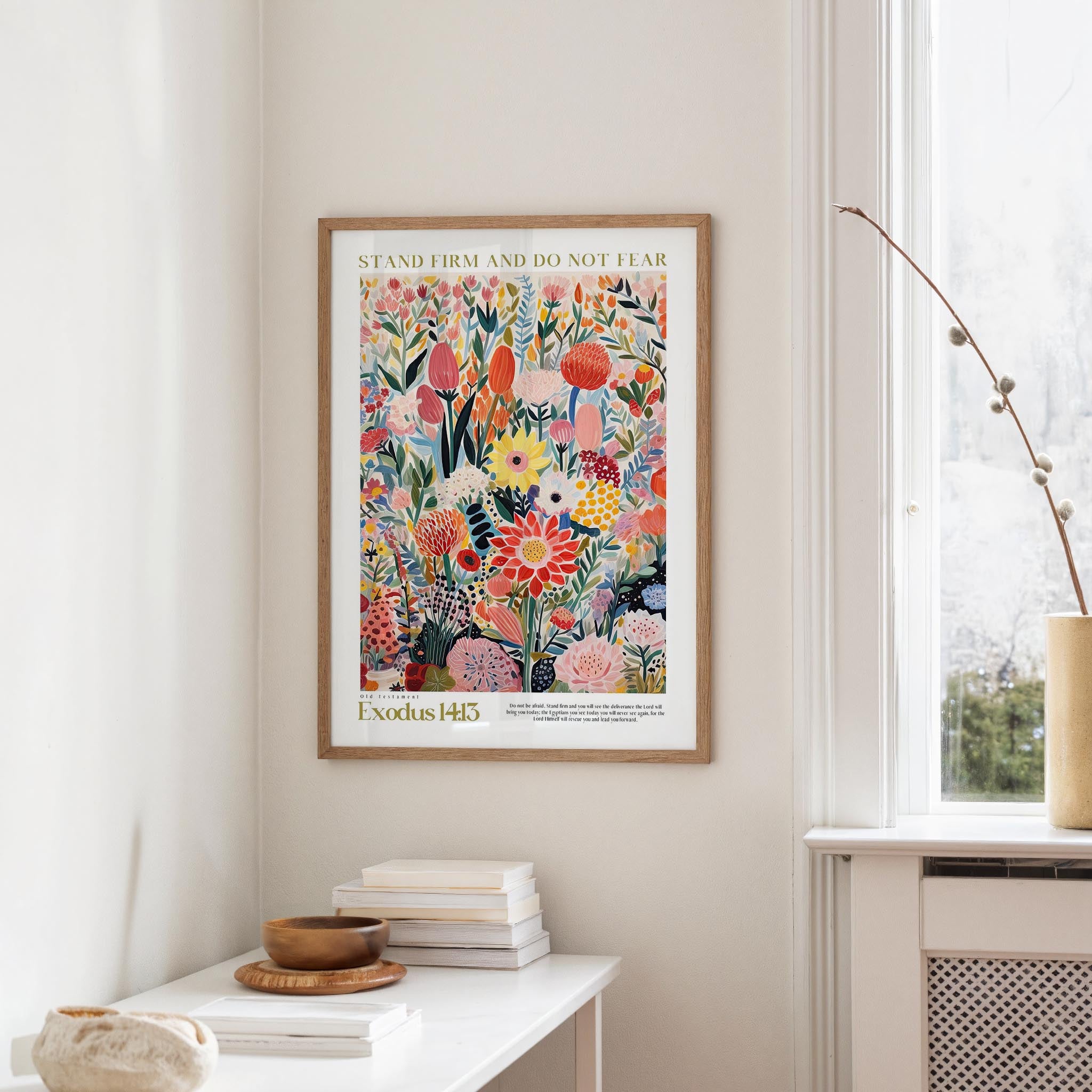 a framed poster of a colorful floral design hanging on a wall, with a stack of books and a vase on a table below it.