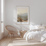 A bedroom with a bed, a woven chair, and a framed poster of a mountain landscape hanging on the wall.