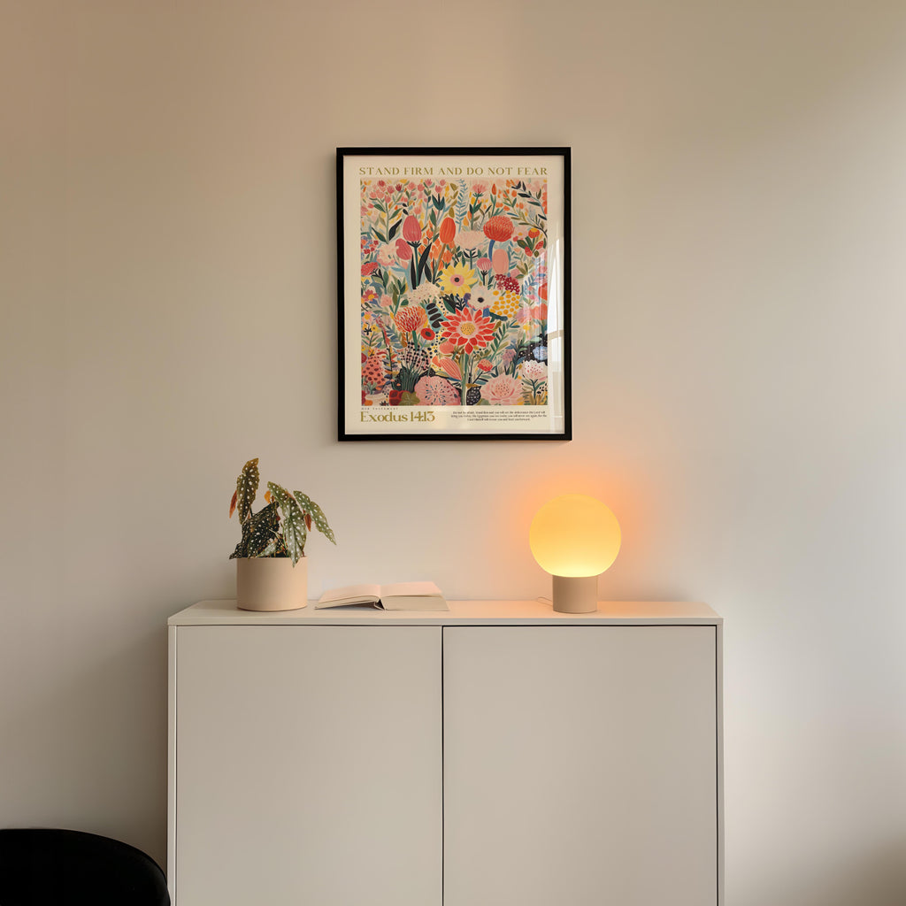 a white cabinet with a potted plant and a lamp on top, set against a white wall. Above the cabinet, there is a framed poster with a floral design.